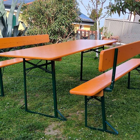 Eine breite Bierzeltgarnitur in der Farbe Ocker mit Rückenlehne und grünem Untergestell steht in einem privaten Garten zwischen Bäumen.